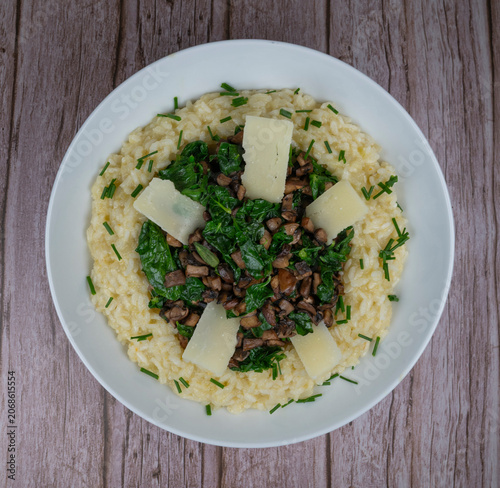 A bowl contains a serving of mushroom and spinach risotto. On top are pieces of Morteau sausage and parmesan cheese. The dish is placed on a wooden surface.