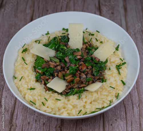 A bowl contains a serving of mushroom and spinach risotto. On top are pieces of Morteau sausage and parmesan cheese. The dish is placed on a wooden surface.