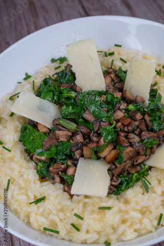 A bowl contains a serving of mushroom and spinach risotto. On top are pieces of Morteau sausage and parmesan cheese. The dish is placed on a wooden surface.