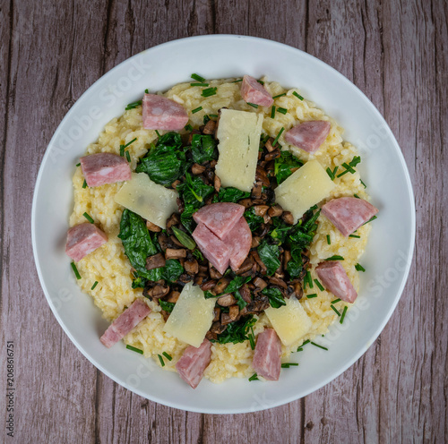 A bowl contains a serving of mushroom and spinach risotto. On top are pieces of Morteau sausage and parmesan cheese. The dish is placed on a wooden surface.