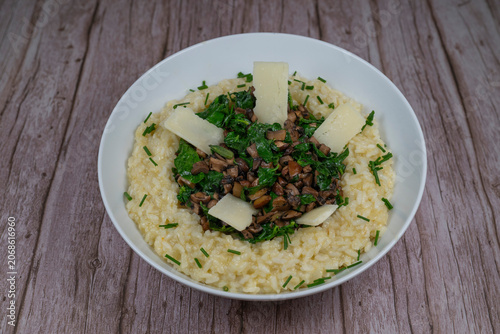 A bowl contains a serving of mushroom and spinach risotto. On top are pieces of Morteau sausage and parmesan cheese. The dish is placed on a wooden surface.