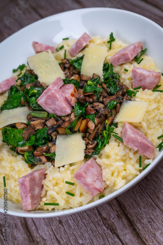 A bowl contains a serving of mushroom and spinach risotto. On top are pieces of Morteau sausage and parmesan cheese. The dish is placed on a wooden surface.