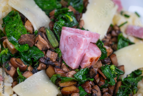 A bowl contains a serving of mushroom and spinach risotto. On top are pieces of Morteau sausage and parmesan cheese. The dish is placed on a wooden surface.