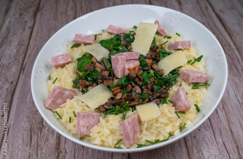 A bowl contains a serving of mushroom and spinach risotto. On top are pieces of Morteau sausage and parmesan cheese. The dish is placed on a wooden surface.