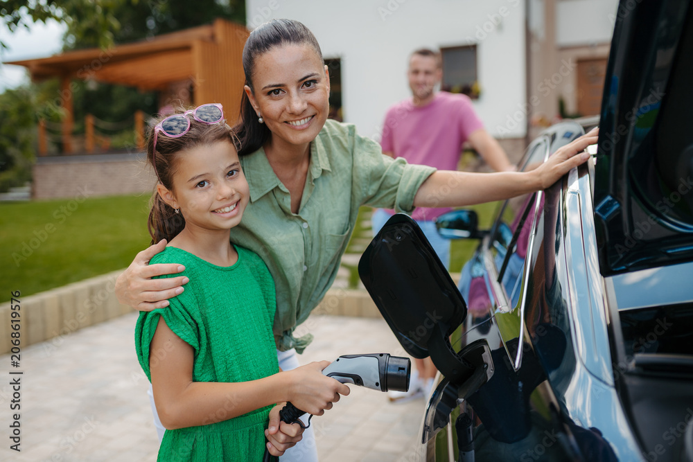 Fototapeta premium Mother and girl charging electric car while family prepares for summer trip.