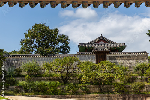 Traditional Korean palace pavilion and historic stone wall on a green terraced hill under a cloudy blue sky.