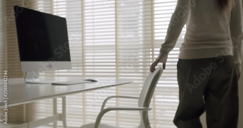 Businesswoman walking into a bright, modern office and pulling out her chair to start working at the desk with a computer. Slow motion shot showing the beginning of a productive workday