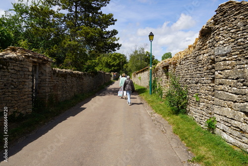 People walk along a street with high old stone walls with doorways as an entrance to the yard in the village of Châteauneuf. Blue sky, June. Burgundy, France. 