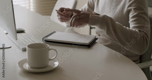 Anonymous woman sitting at a modern white desk next to a computer, opening a spiral notebook and beginning to write with a pencil, a cup of coffee resting nearby in a bright minimalist office