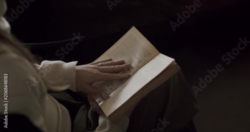 Close-up view of a woman sitting in a dark room and reading an old book with yellowed pages, her fingers gently turning the pages as she becomes engrossed in the story, enjoying a quiet moment