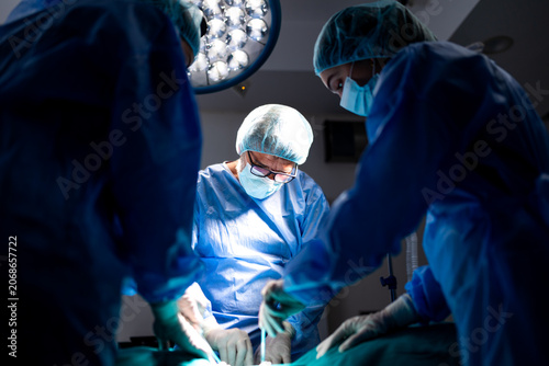 Intense focus of a surgical team performing an operation under bright overhead lights in a modern hospital operating room. Surgeon and assistants in blue scrubs work with precision on a patient.