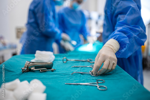 Medical professional in blue surgical scrubs and sterile gloves reaches for stainless steel surgical instruments on a tray in a sterile operating room environment during a planned surgical procedure.