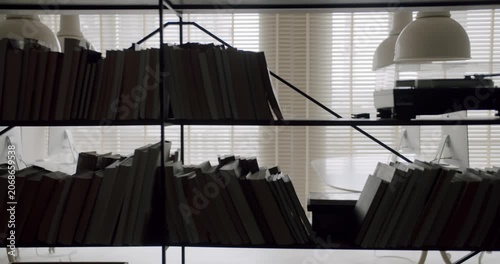 Dark modern bookshelf silhouette filled with numerous books and a vinyl record player standing against a window with closed blinds, revealing a sleek and stylish contemporary interior