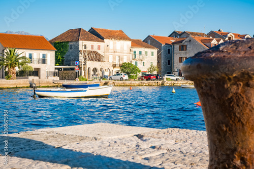 Mooring bollard in Tivat, Montenegro. A small white boat is anchored in the blue water. Colorful buildings line the shore under a clear sky.