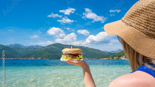 A young girl with long blonde hair holds a burger in front of a clear blue sea and mountains under a bright sky.