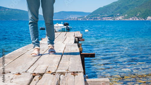 A woman walks along a wooden pier over clear blue water in Tivat, Montenegro. In the background, green hills and distant buildings are visible.