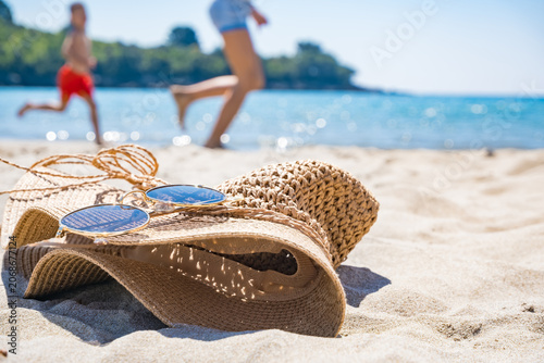 A straw hat and sunglasses lie on the sandy beach. Two people, a young boy in red shorts and a woman in shorts, run along the shore in the background.
