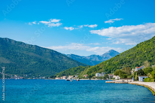 Scenic view of Tivat, Montenegro. Clear blue water, green hills, and mountains under a bright sky. A peaceful coastal landscape with a serene atmosphere.