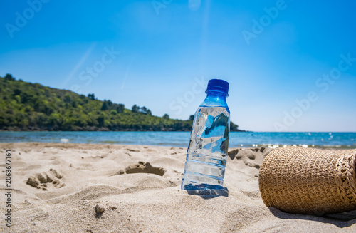 A clear water bottle stands on golden sand at the beach. A straw hat lies beside it. Ocean waves gently lap the shore under a bright blue sky.
