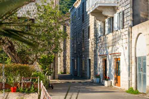 A narrow street in Tivat, Montenegro, lined with stone buildings. Green plants and flowers add color to the scene. Bright sunlight illuminates the area.