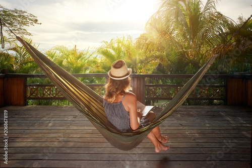 Young woman in straw hat relaxing in hammock on wooden terrace, reading book in tropical resort. Summer vacation concept.