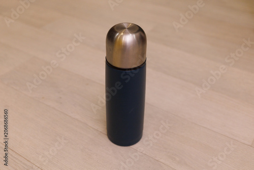 A thermal bottle stands upright on a light wooden floor in a room