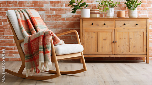 Cozy rocking chair with plaid blanket near wooden cabinet and brick wall in warm room