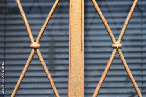 Slika na platnu Close-up of a vintage metallic safety grill with criss-cross pattern and rusty golden texture over a window with closed blue blinds