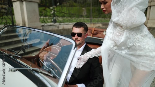 Groom at wheel of vintage convertible as bride leans on car at grand estate