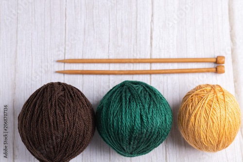 Three yarn balls in brown, green and mustard colors with wooden knitting needles on light surface. Horizontal minimalist photo with neutral background and copy space