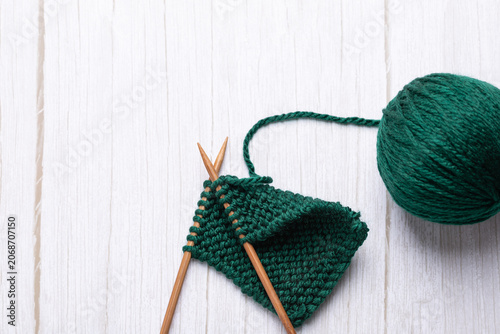 Top view of a green yarn ball with knitting needles inserted into garter stitch fabric on a light wooden table. Minimalist vertical image with copy space. Concept of knitting craft and slow living