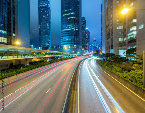 A long exposure photograph captures the vibrant streaks of light from moving vehicles along a wide city avenue, framed by towering skyscrapers under a twilight sky.