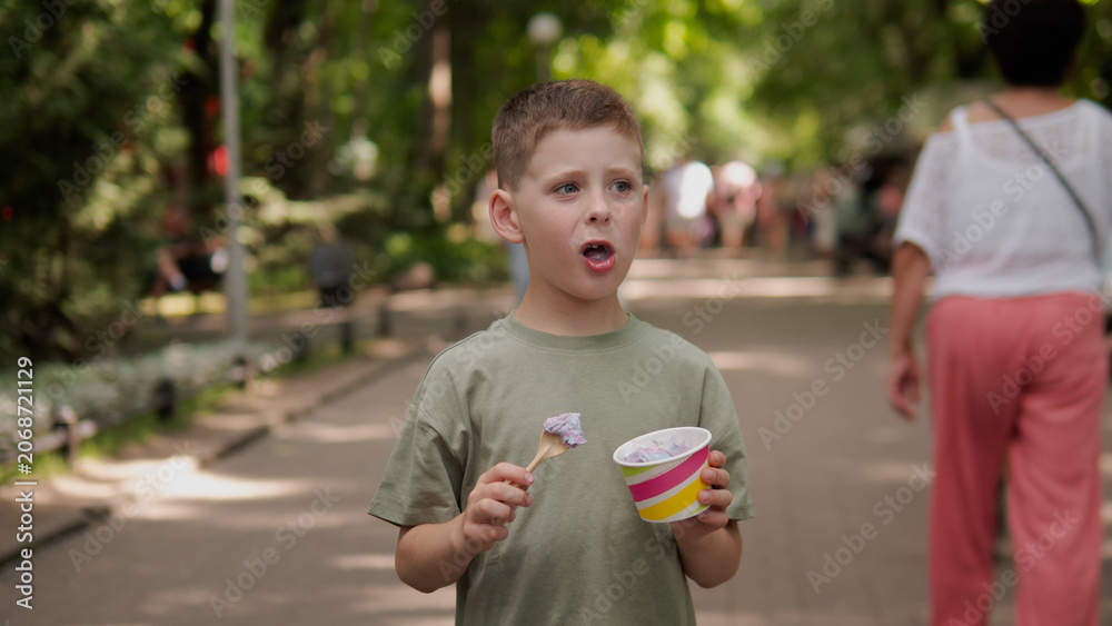 Naklejka premium A young boy stands on a sunlit park path holding a cup of ice cream, with a surprised expression and a colorful cup in hand