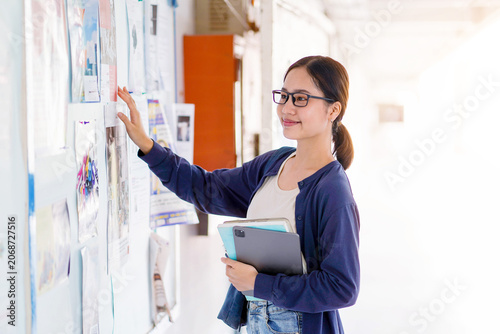 Obraz na plátně Happiness teenage Asian girl, dressed in casual clothes, holds a tablet and finding part-time job on school bulletin board in her school building