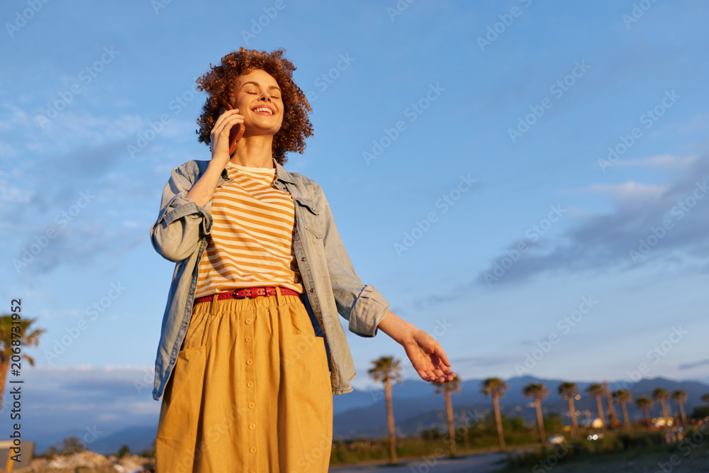 Obraz premium Young woman outdoors enjoying a sunny day, casual fashion with a denim jacket over a striped tee and a mustard skirt, a vibrant lifestyle portrait on an open coastal road.