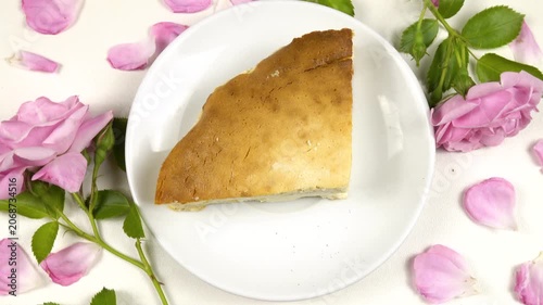 A white plate with a piece of homemade cottage cheese pie on it stands on a white table among roses and petals, top view