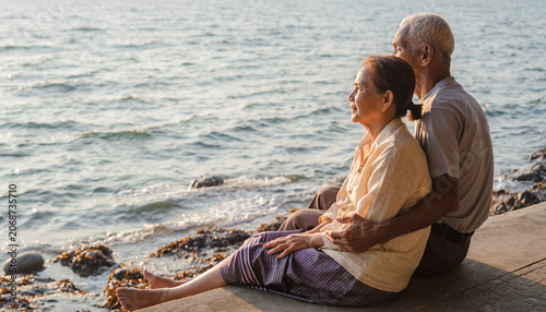 An elderly couple sits closely together on a concrete edge overlooking the gently rippling water of the sea or ocean.