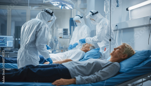Medical staff in full protective gear attend to patients lying on hospital beds in a brightly lit, modern intensive care unit setting.