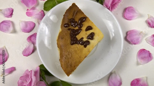 White plate with a cottage cheese pie sits on a white table, among roses and petals.  A slice of homemade cheesecake is being covering with chocolate icing, top view