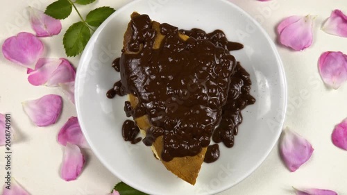 A white plate with a piece of homemade cottage cheese pie with chocolate icing on it stands on a white table among roses and petals, top view