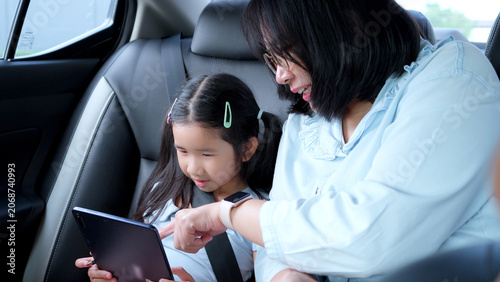 Photos Asian mother teaching her cute little daughter with a digital tablet in the back seat of a car during a family road trip, parenting and safety travel concept