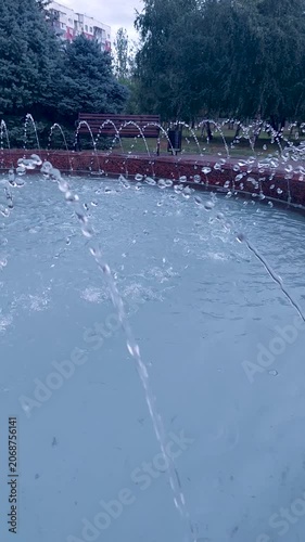 Close-up of fountain water jets and droplets falling into a pool on a gray rainy day in a park with a bench in the background
