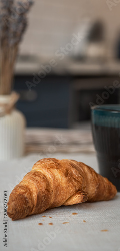 Golden buttery croissant on a table with coffee cup in the background. Cozy breakfast atmosphere with shallow depth of field and copy space.