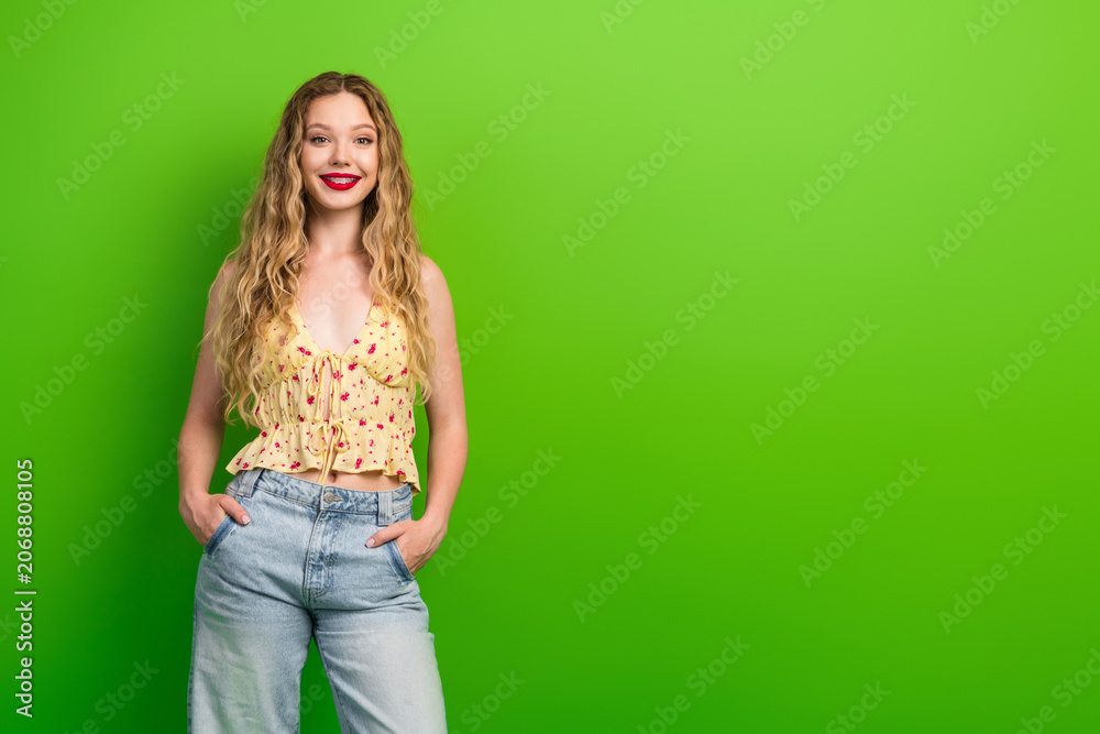 Obraz premium Glamorous young woman with blonde hair posing in floral top and jeans against a vivid green background