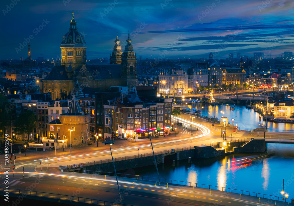Obraz premium Stunning blue hour panoramic view of Amsterdam city centre with Basilica of Saint Nicholas and canals, illuminated by glowing city lights under a dramatic twilight sky, Netherlands