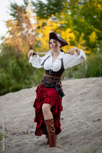 Schilderij op canvas Full-length portrait of a young woman dressed in a pirate costume posing with a saber in her hands while standing against the backdrop of the shore