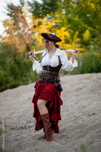 Schilderij op canvas Full-length portrait of a young woman dressed in a pirate costume posing with a saber in her hands while standing against the backdrop of the shore