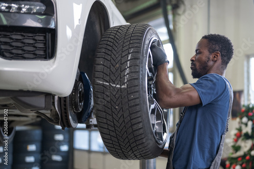 African American mechanic securely installing a car wheel on the vehicle raised by a hydraulic lift in a service garage.