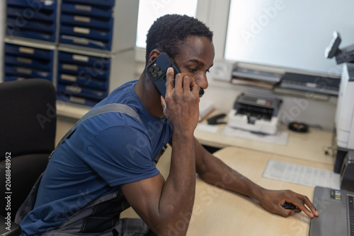 A male auto mechanic is on the phone with a customer while working at a computer in an automotive repair workshop.