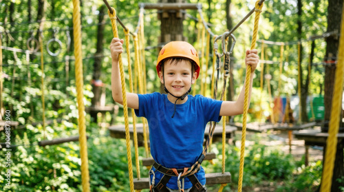 Young boy in blue shirt and orange helmet smiles while holding onto ropes in a tree climbing adventure park surrounded by greenery